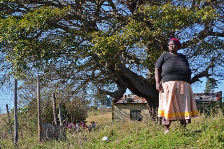 Activist Duduzile Dludla of Makhasaneni, standing in front of her home and an umbombe or forest strangler-fig (Ficus natalensis)