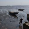 Manoka, Cameroon: solitary fisher with a fish in their right hand, stepping away from three canoes beached on a shimmering mudflat, to a concrete jetty in the bottom right. Image by Christophe Nyemeck Beat
