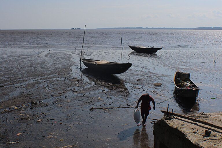 Manoka, Cameroon: solitary fisher with a fish in their right hand, stepping away from three canoes beached on a shimmering mudflat, to a concrete jetty in the bottom right. Image by Christophe Nyemeck Beat