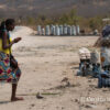 A woman in a yellow top and bright multicoloured wrapper, smiling as she walks across sandy verge of a paved road to where blue and white bags of dark black charcoal have been set on the ground near Praça das Mangueiras, in southwestern Angola. In the background, more charcoal in tall white sacks. Image by Carsten Ten Brink via Flickr (BY NC-ND 2.0)